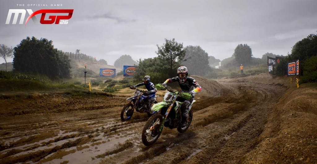 Due motociclisti sfrecciano su un percorso fangoso in un evento di motocross, con alberi e bandiere visibili sullo sfondo.